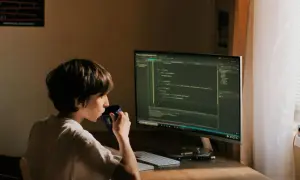 Un jeune garçon boit dans une tasse bleue devant un écran d'ordinateur affichant du code. Il est un développeur indépendant en herbe.