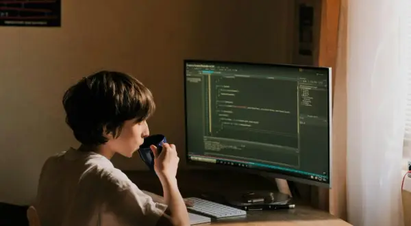 Un jeune garçon boit dans une tasse bleue devant un écran d'ordinateur affichant du code. Il est un développeur indépendant en herbe.