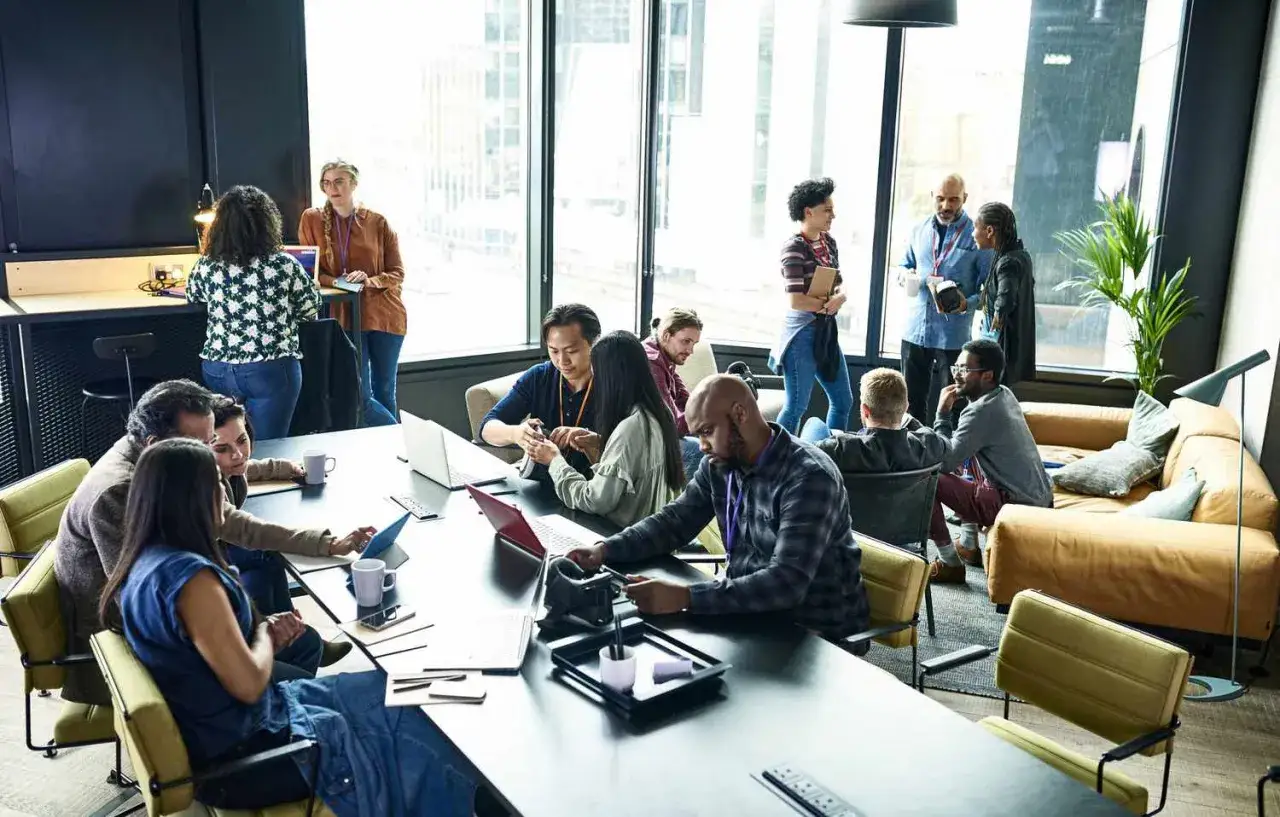 Un formateur web anime une session de travail collaborative dans un espace moderne. Des participants échangent autour d'une table, certains sur leurs ordinateurs.