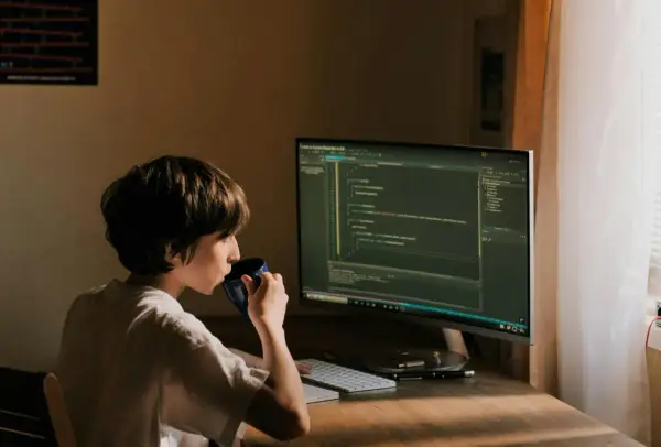 Un jeune garçon boit dans une tasse bleue devant un écran d'ordinateur affichant du code. Il est un développeur indépendant en herbe.
